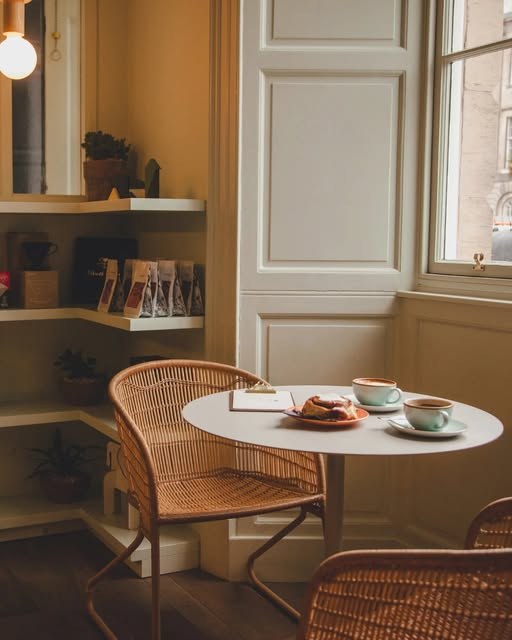 A cozy corner table at Mayvn with two coffees and pastries, a wicker chair and retail bean bags on a shelf behind