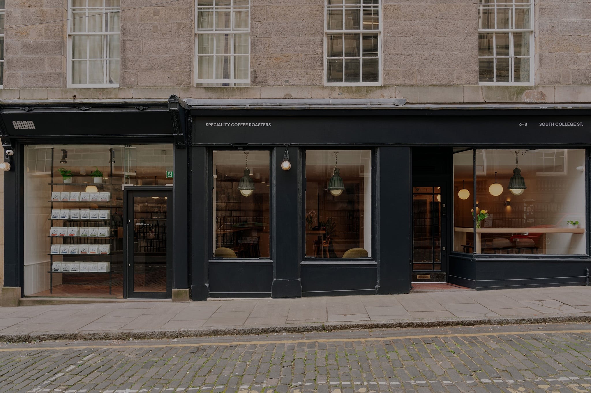 Origin Coffee's black shopfront on South College Street with ORIGIN and SPECIALITY COFFEE ROASTERS signage above large windows showing warm pendant lighting and a retail shelf of coffee bags inside