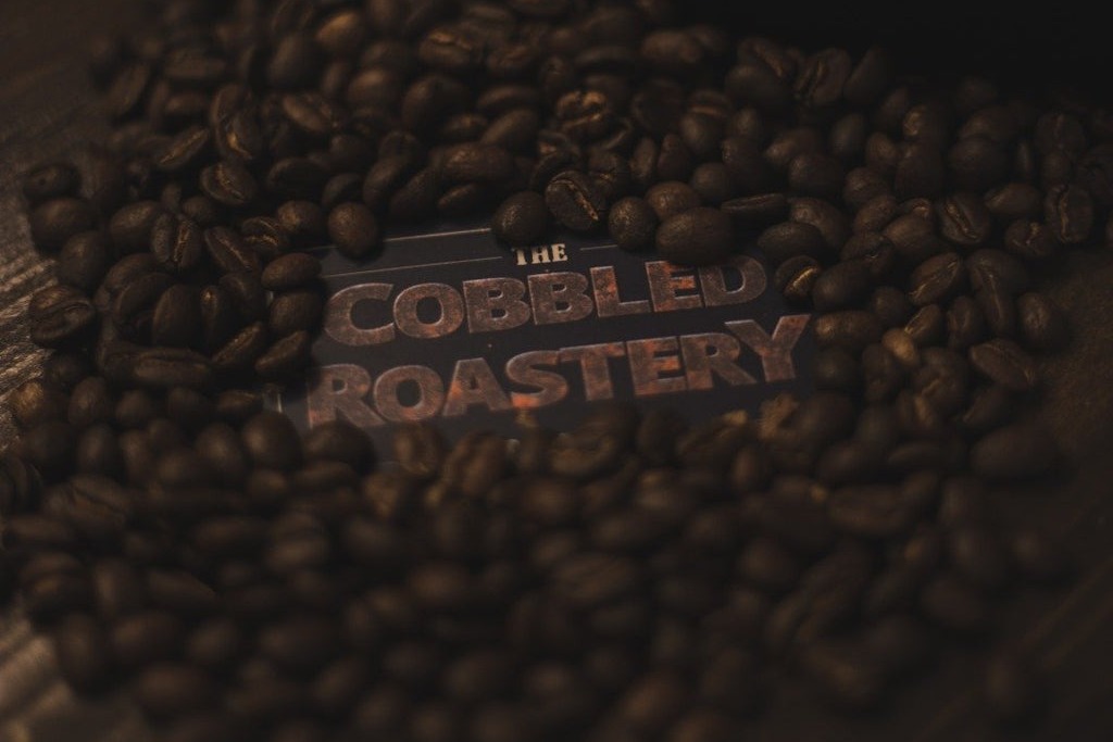 A dark branded card reading THE COBBLED ROASTERY nestled among freshly roasted coffee beans spilling across a wooden counter