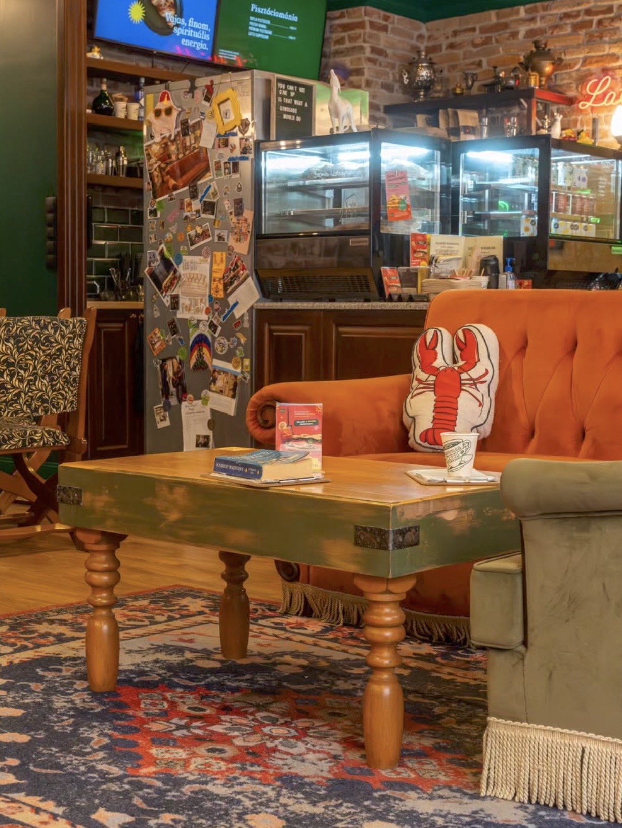 The Friends themed seating area at Central Park Eger, with an orange velvet couch, a lobster cushion, and a to go coffee cup on the table
