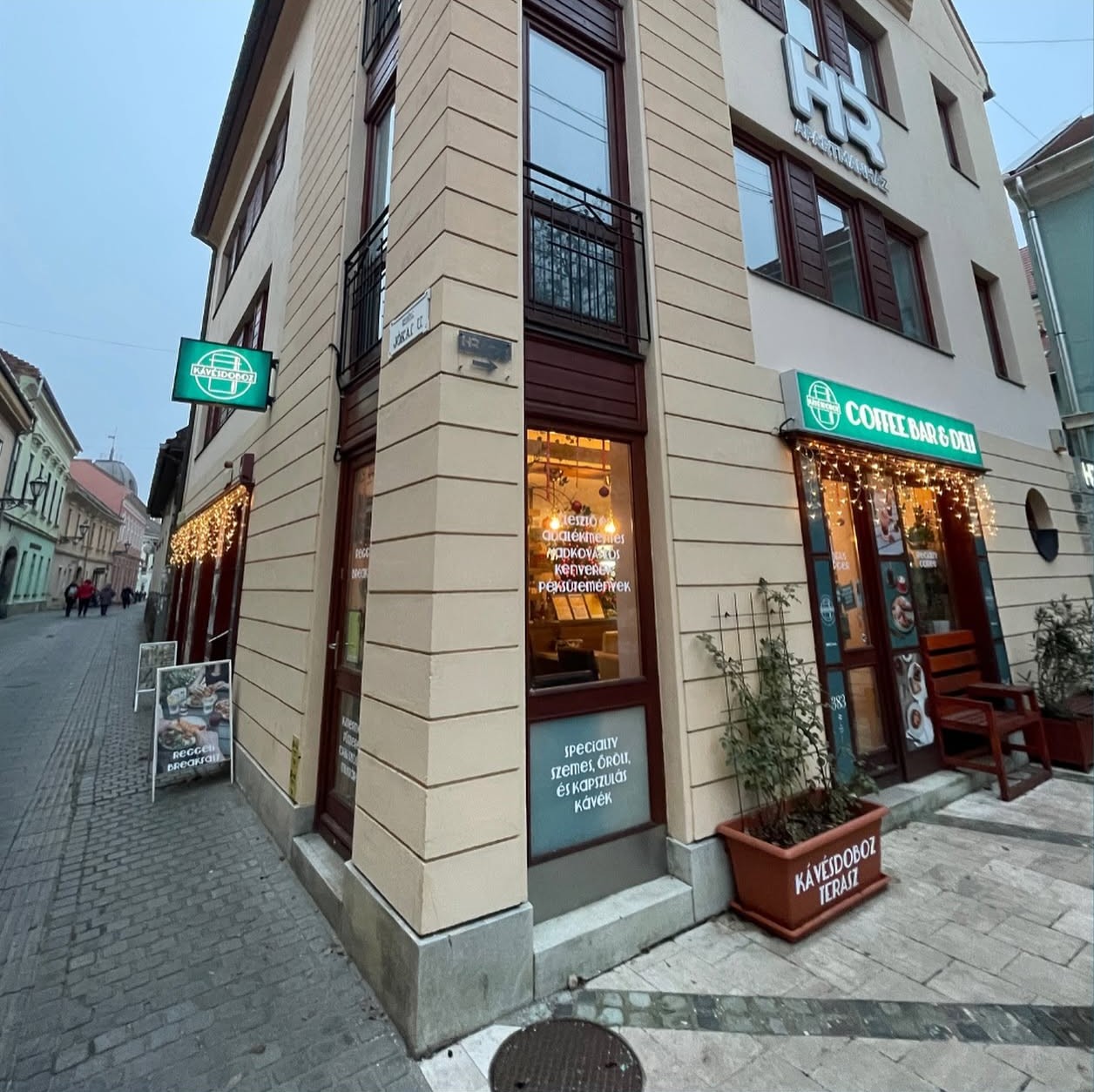 The corner storefront of Kávésdoboz Coffee Bar & Deli in Eger, with a lit green sign and awning reading Coffee Bar and Deli