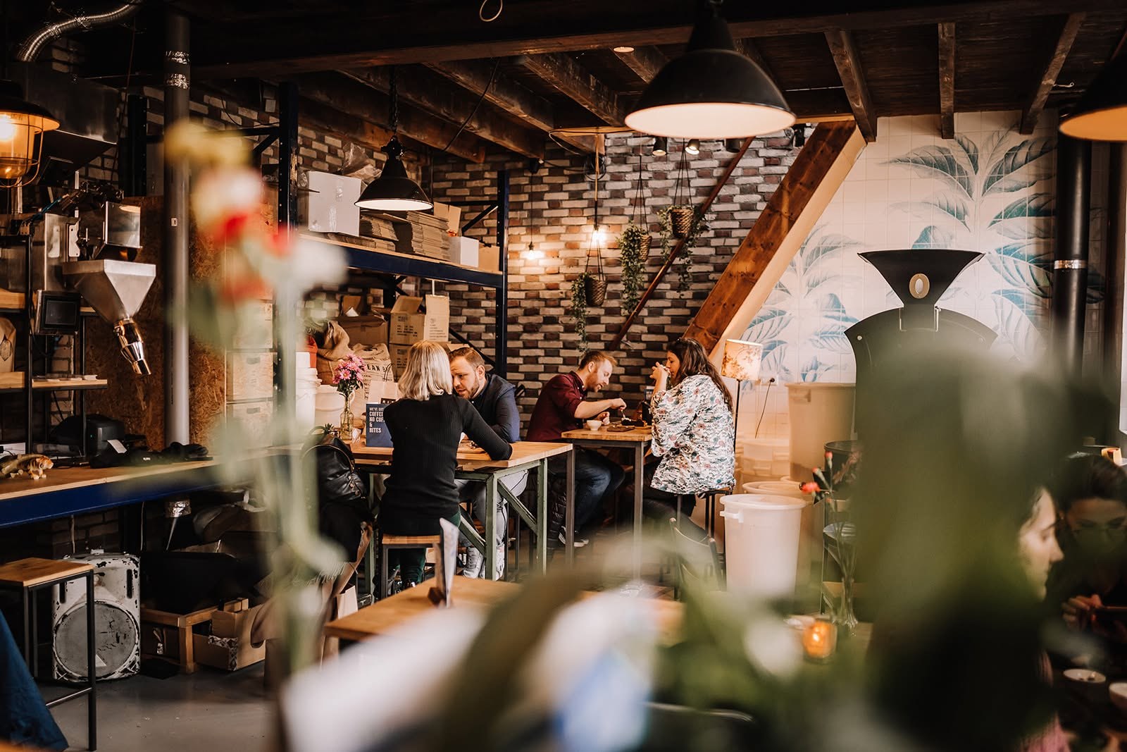 The industrial roastery cafe interior with exposed brick, hanging plants and a large roaster against a leaf mural, customers seated at communal tables