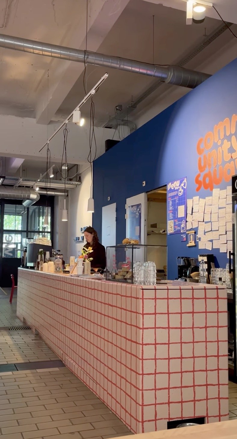 The red gridded tile counter beneath the Community Square wall lettering, staff working the espresso machine