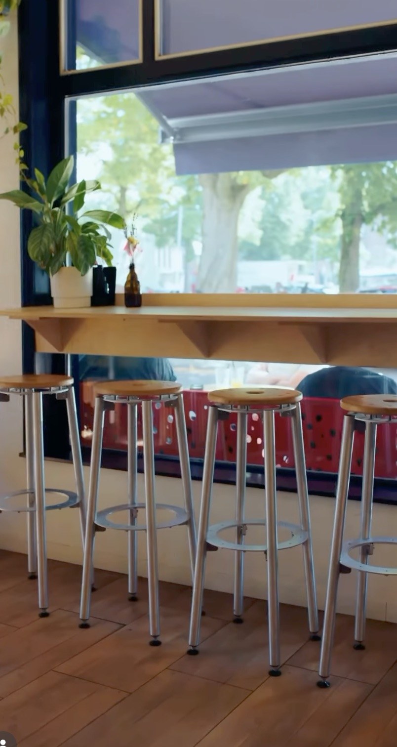 Wooden bar stools along a tall window counter with a potted plant, street trees visible outside
