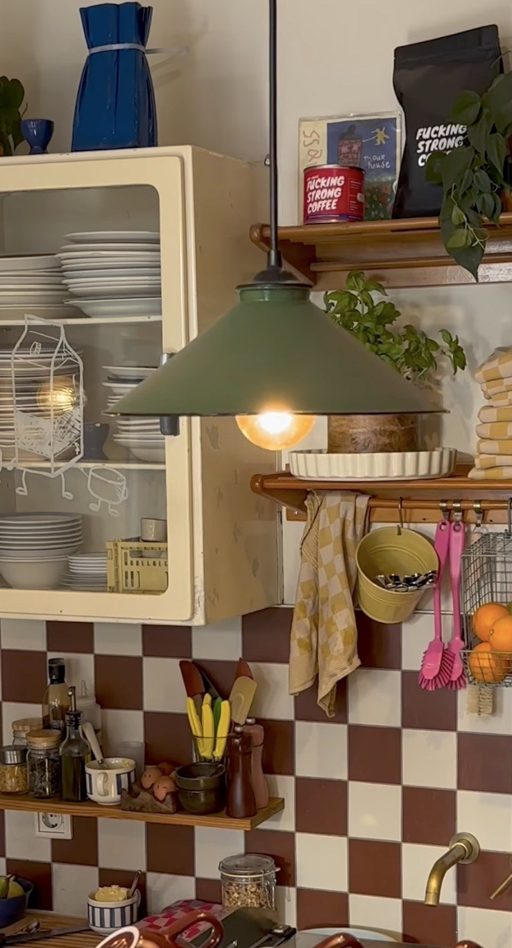 An open crockery cabinet and a green pendant lamp above a shelf holding a retail coffee bag and tin, checkerboard tile behind