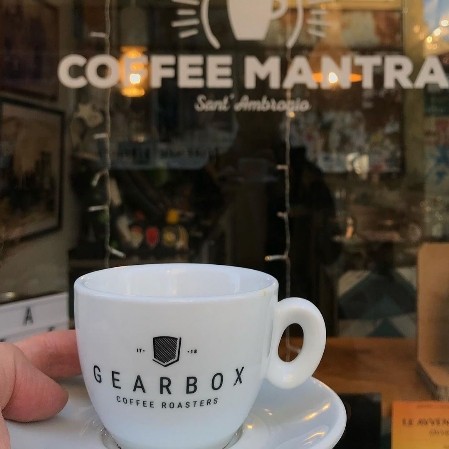 A Gearbox Coffee Roasters cup held in front of the Coffee Mantra Sant'Ambrogio storefront window