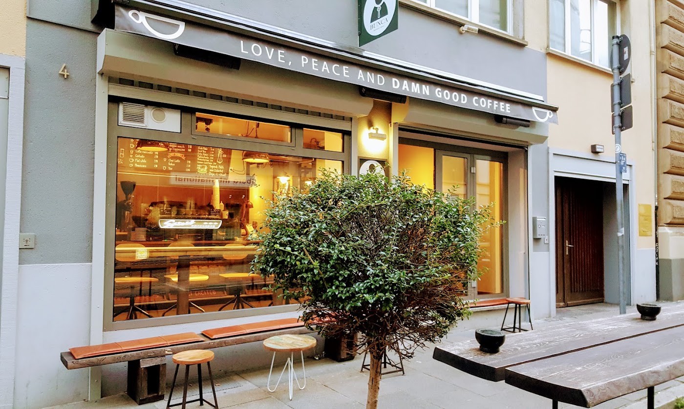 BUNCA's narrow storefront on Kirchnerstraße at dusk, its lit counter visible through the window behind a small potted tree and outdoor benches