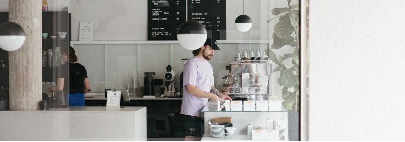 A barista in a pale lilac shirt pulling shots at the espresso machine, menu board and pendant lamps above the counter