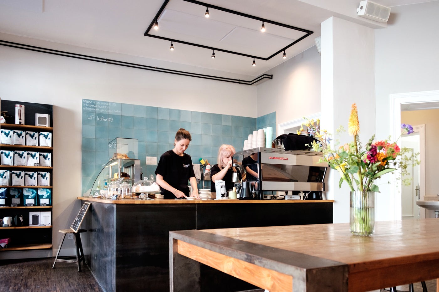 Two baristas working the tiled espresso counter at Hoppenworth & Ploch's Nordend cafe, retail coffee bags on shelving beside a vase of fresh flowers on the communal table