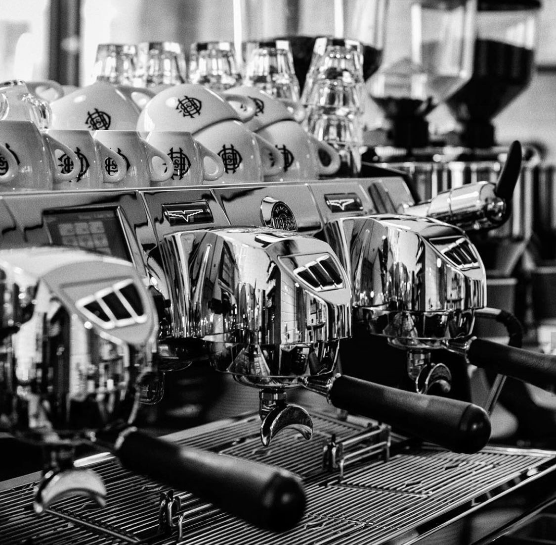 Rows of branded espresso cups lined up behind a polished three group espresso machine at The Holy Cross Brewing Society