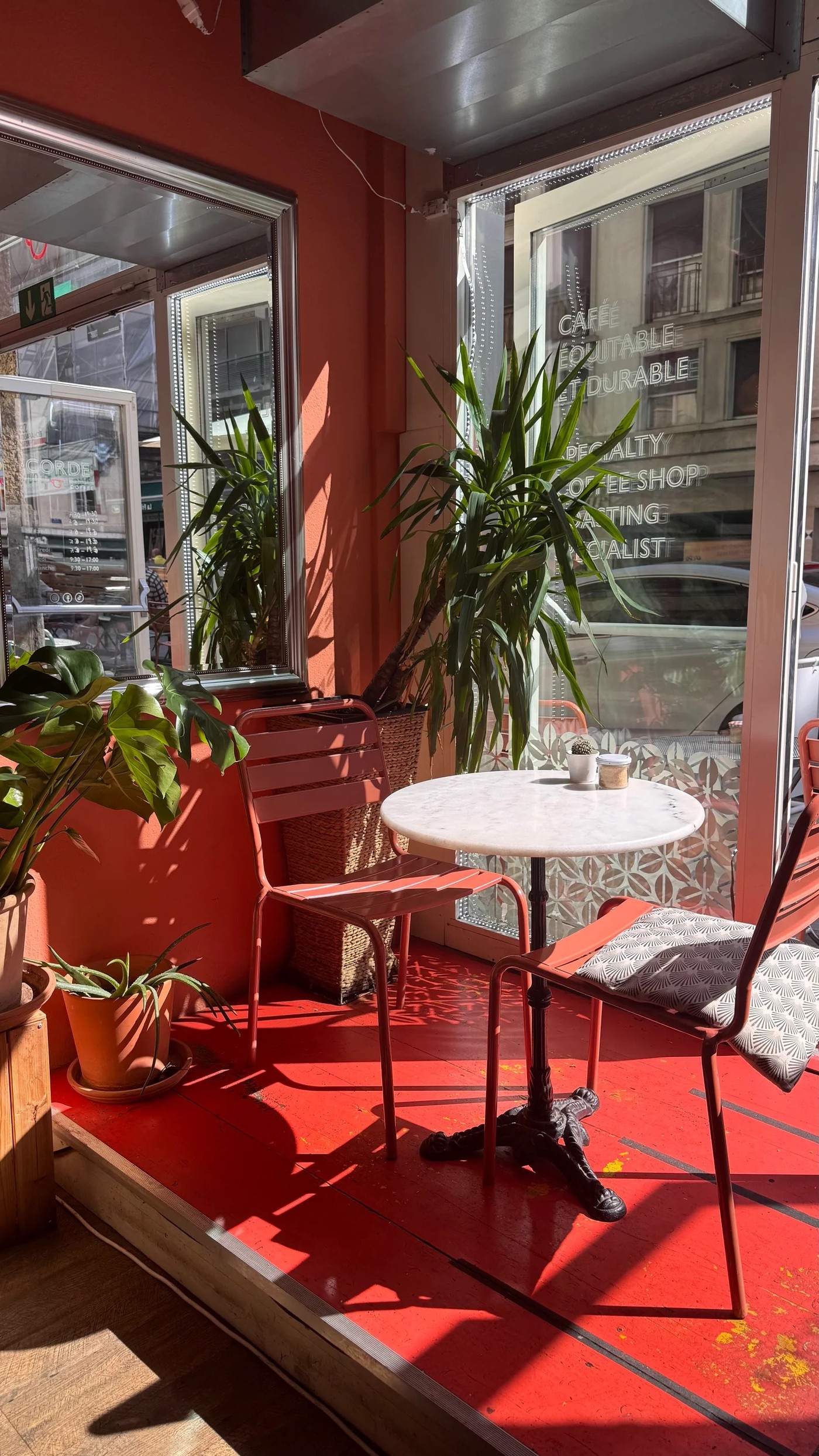 A red toned corner seating nook at Corde Coffee with potted plants, a marble bistro table, and fair trade specialty coffee lettering on the window