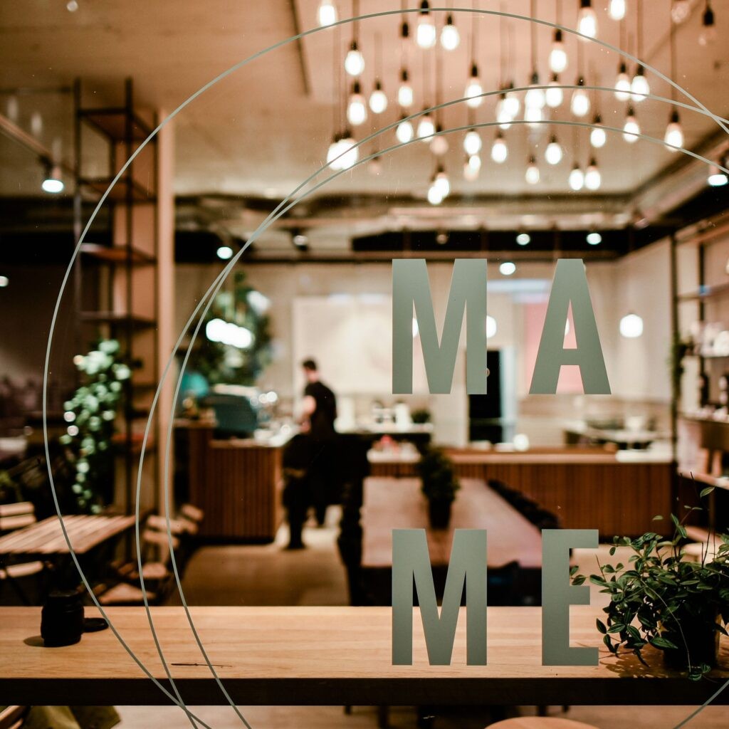 The MAME logo lettering on the storefront window, with the warmly lit interior and string lights visible behind