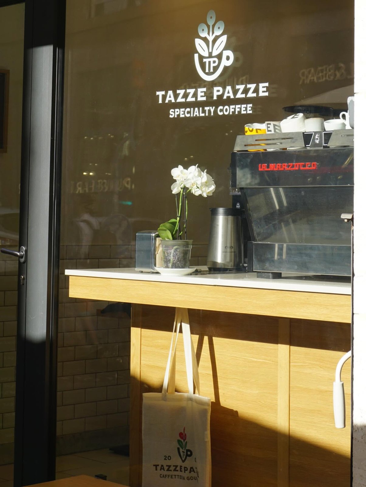 The window at Tazze Pazze with its logo etched on the glass above a La Marzocco espresso machine and a branded tote bag