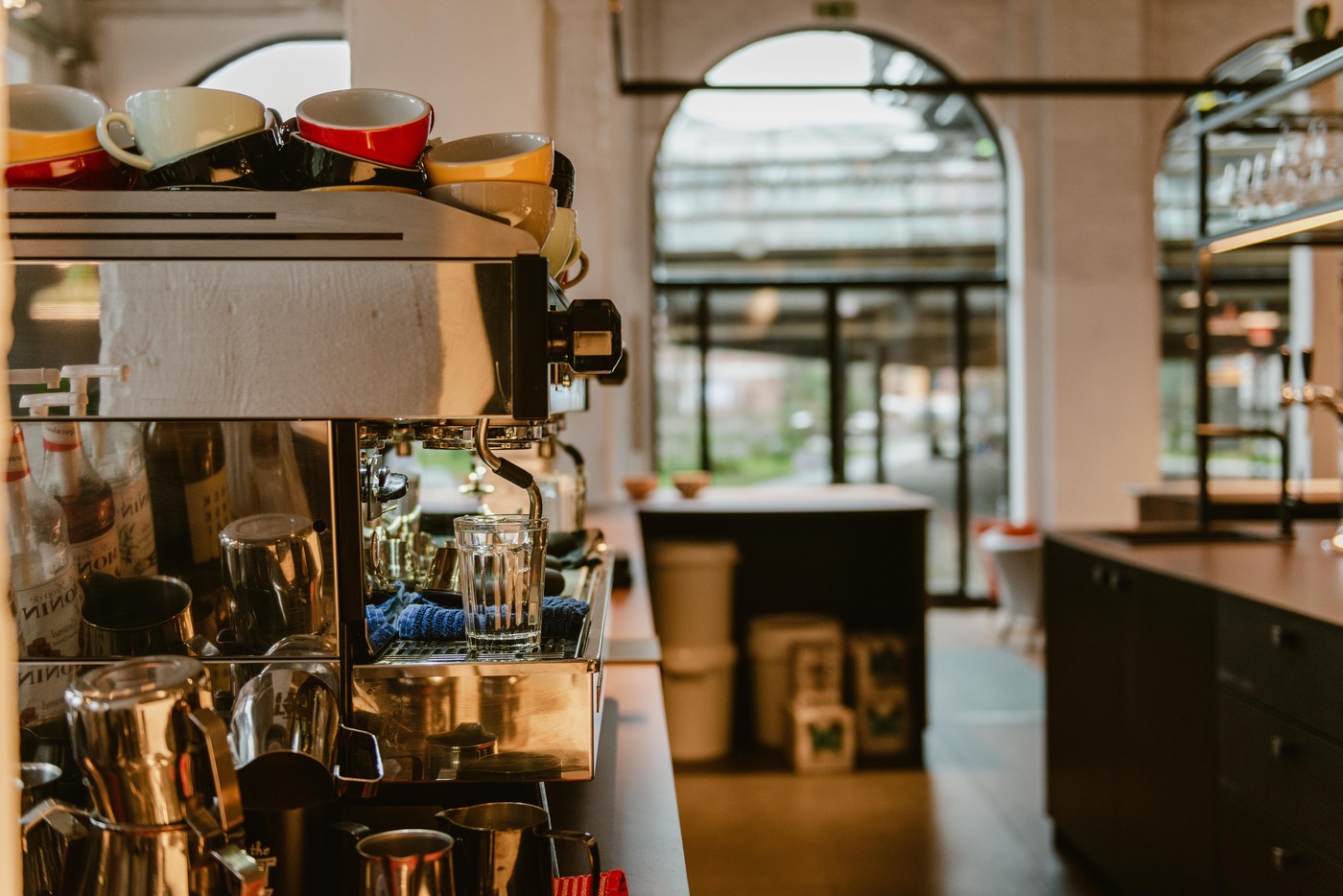 A row of colorful cups stacked on an espresso machine beside a water glass, with an arched window looking onto the street
