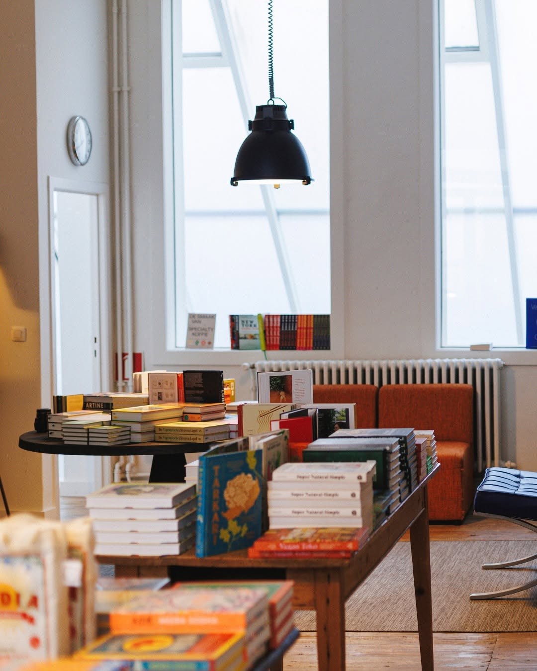 A table stacked with books beside orange armchairs and tall windows inside WAY's Dok Noord space