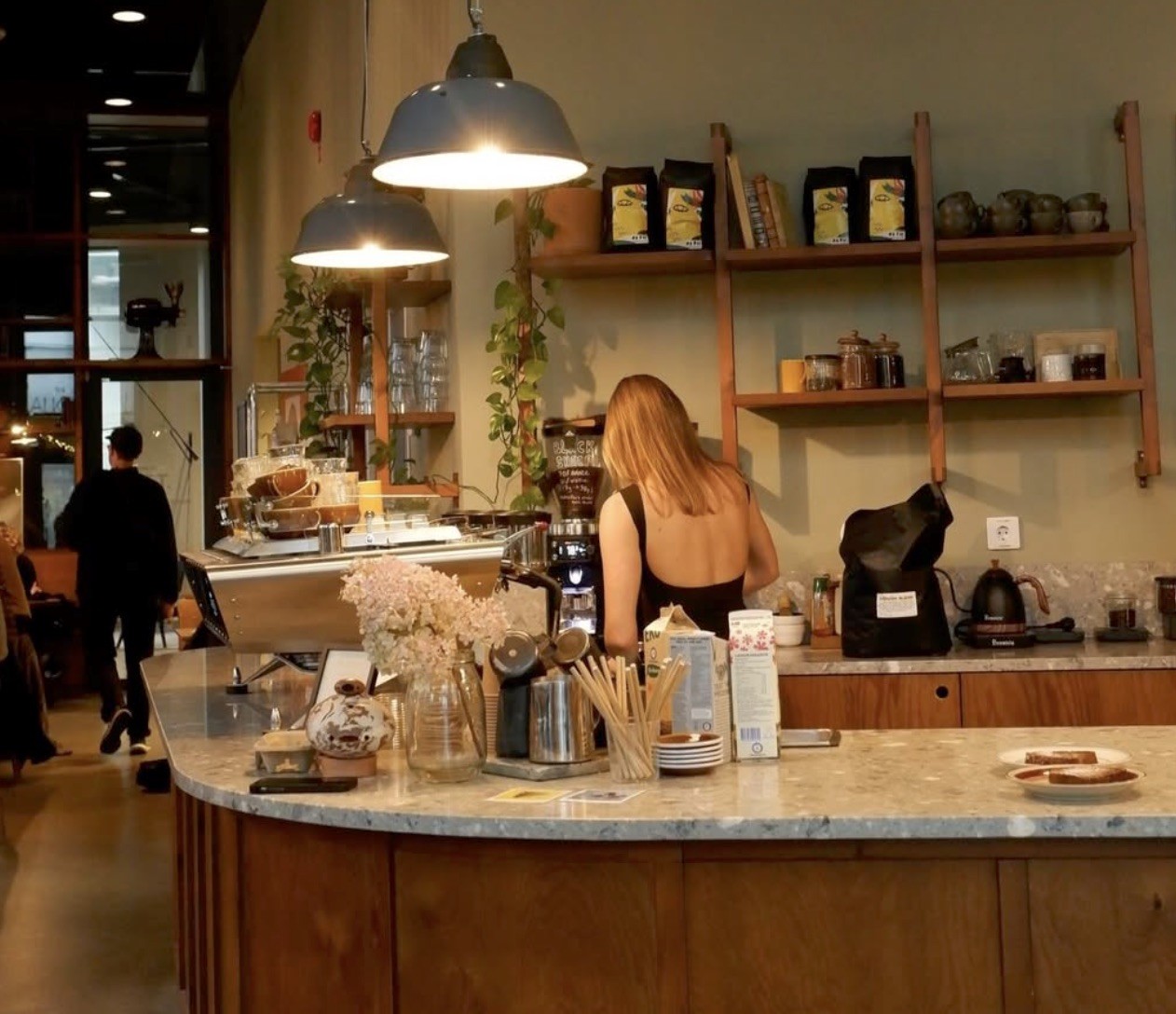 A barista working behind the curved marble counter beneath open wood shelves of retail bags at Alkemisten