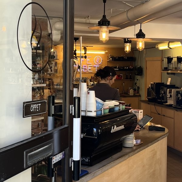 A black La Marzocco espresso machine seen through the front window of Kaffelabbet with a neon sign glowing behind the counter