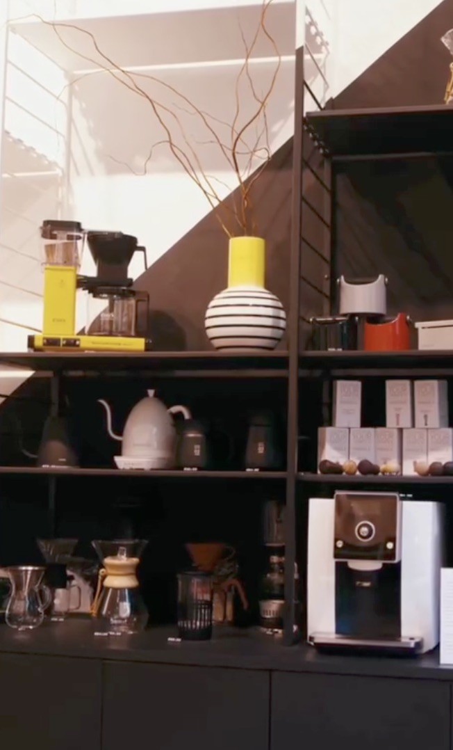 A shelving unit at Buna Coffee Roasters lined with brewing gear, including a Moccamaster, pour over kettles, Chemex, and a bean dispenser