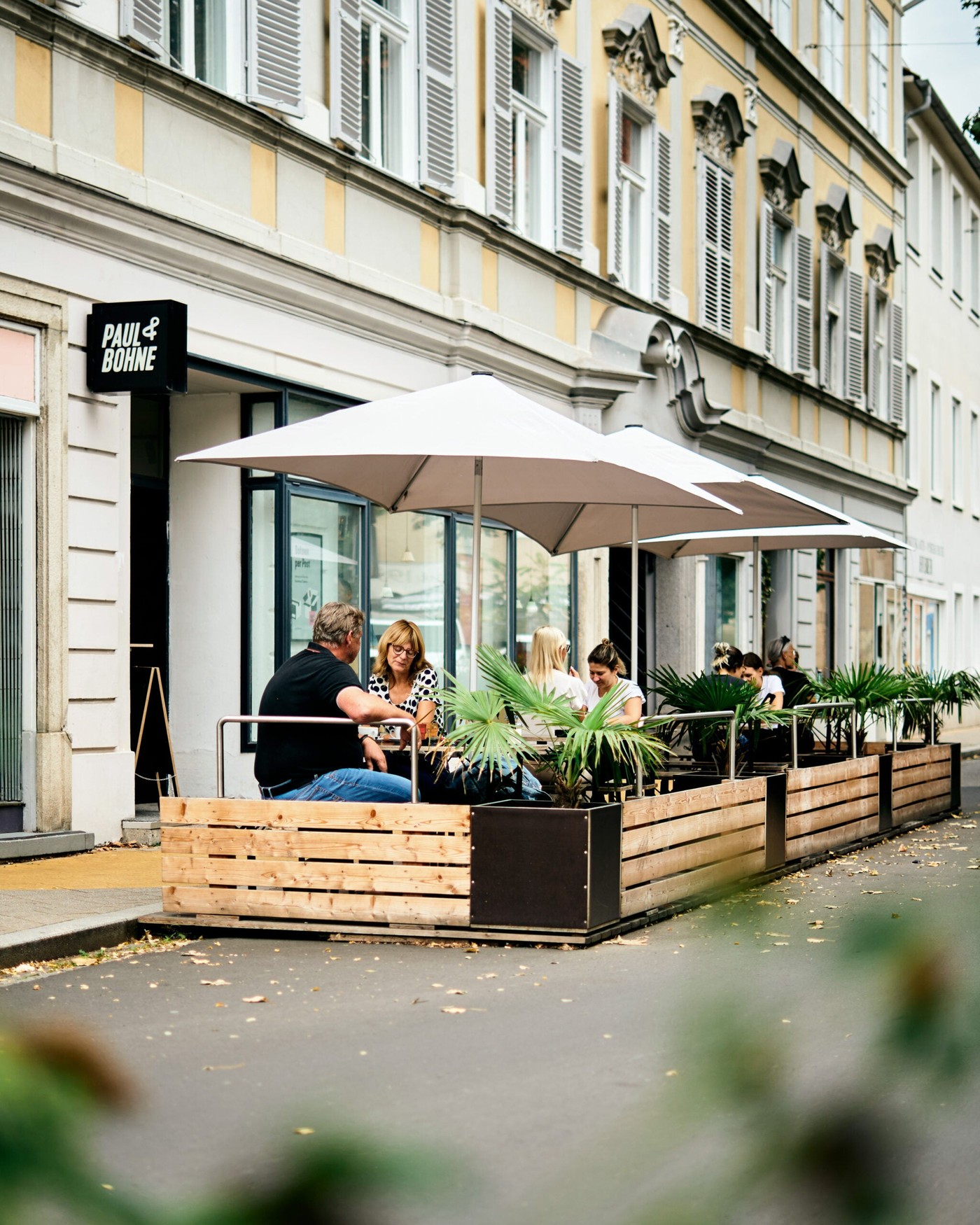 Sidewalk terrace outside Paul & Bohne with the shop's sign above the door and customers seated at wooden planter-lined tables under umbrellas
