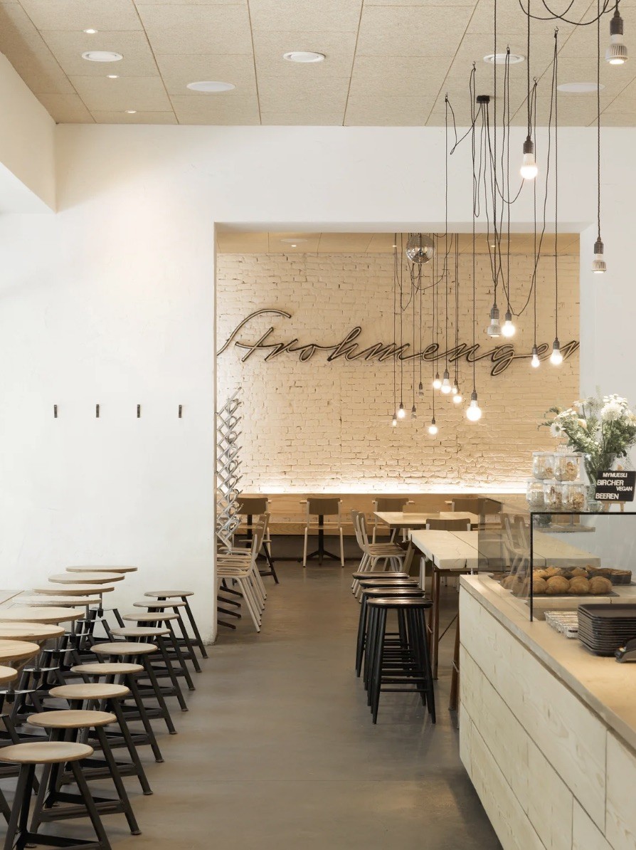 The interior of Tribeka with high stools and communal tables, a bakery counter with pastries, and a lit up cursive sign on the exposed brick wall behind