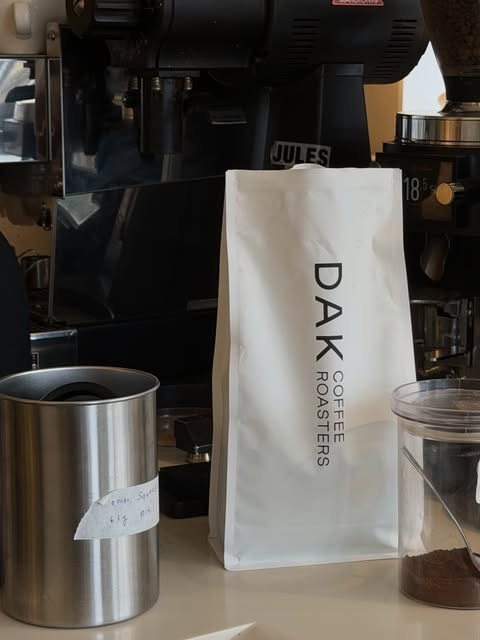 A white DAK Coffee Roasters bag beside the espresso machine and grinder on the counter