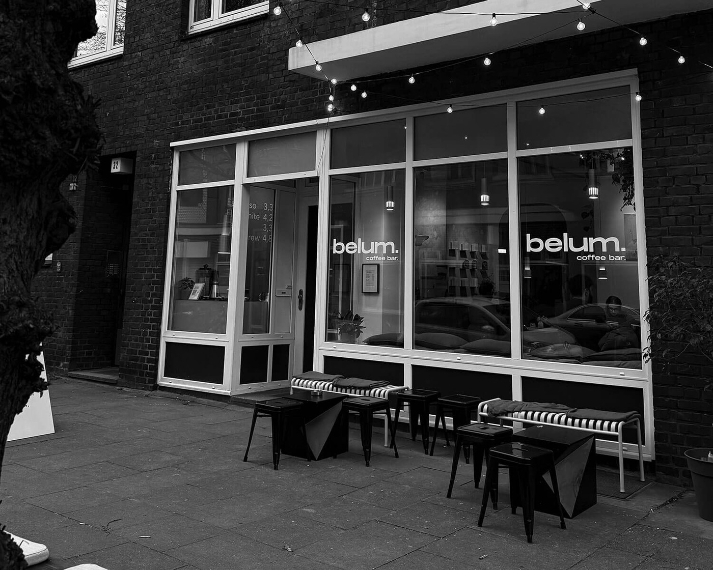 The black and white storefront of belum. coffee bar at dusk, with string lights over an outdoor seating area of stools and low tables