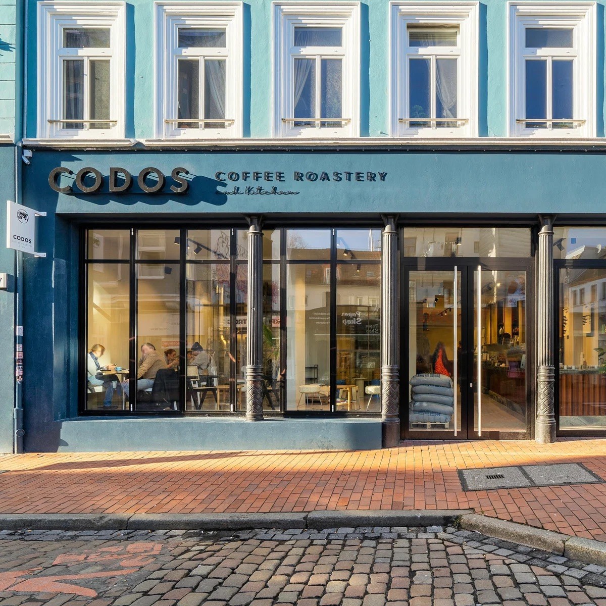The teal storefront of CODOS Coffee Roastery and Kitchen, with ornate columns framing the entrance and customers seated at tables inside