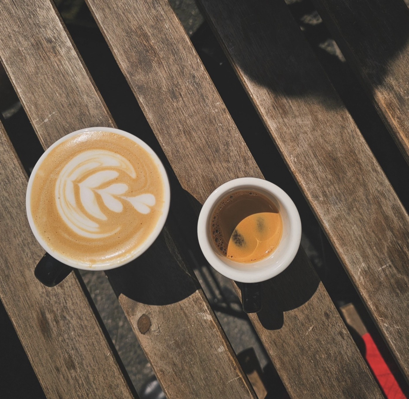 A cappuccino with leaf latte art beside a macchiato in a white cup, shot from above on a wooden bench at Rams Roasters
