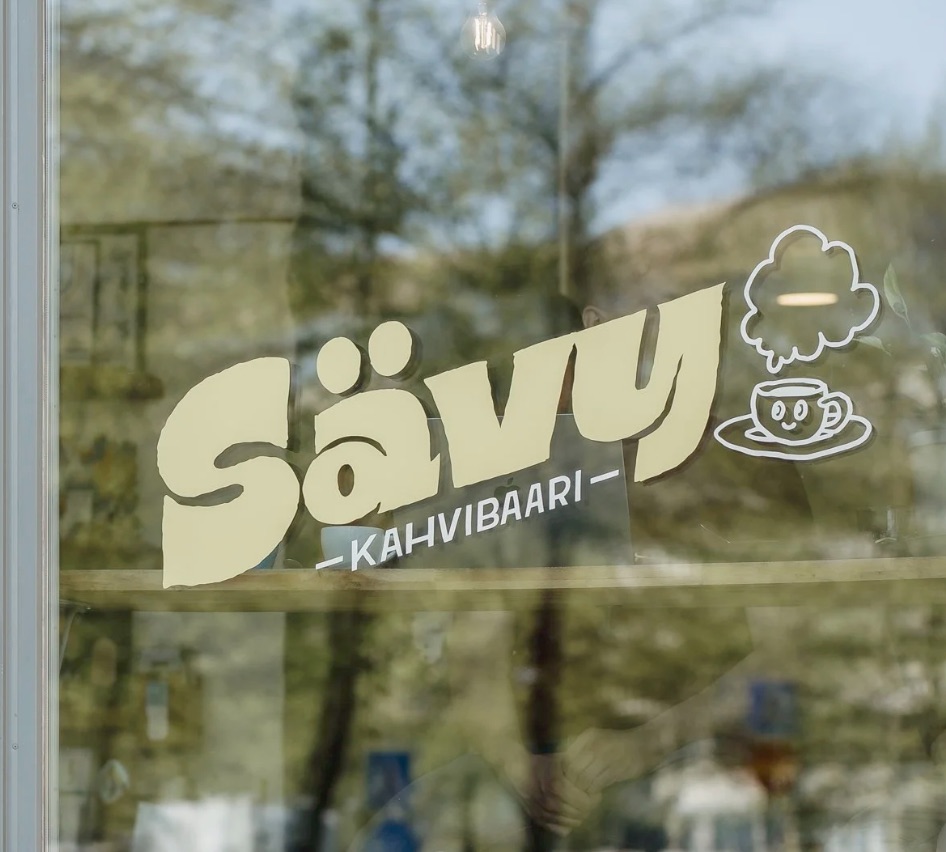 Sävy Kahvibaari's cream logo and a smiling steaming-cup illustration on the cafe's street-facing window, with trees reflected in the glass
