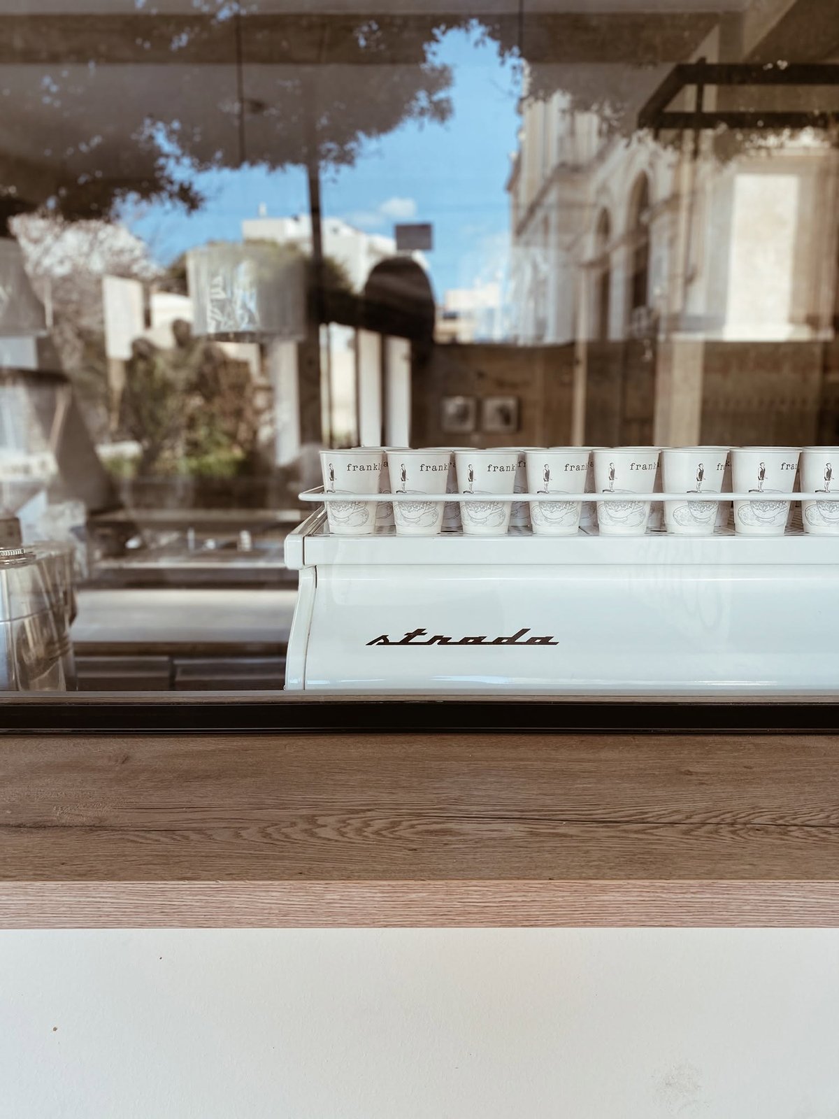 Frankly branded takeaway cups lined up on the La Marzocco Strada espresso machine, with the storefront reflected in the window