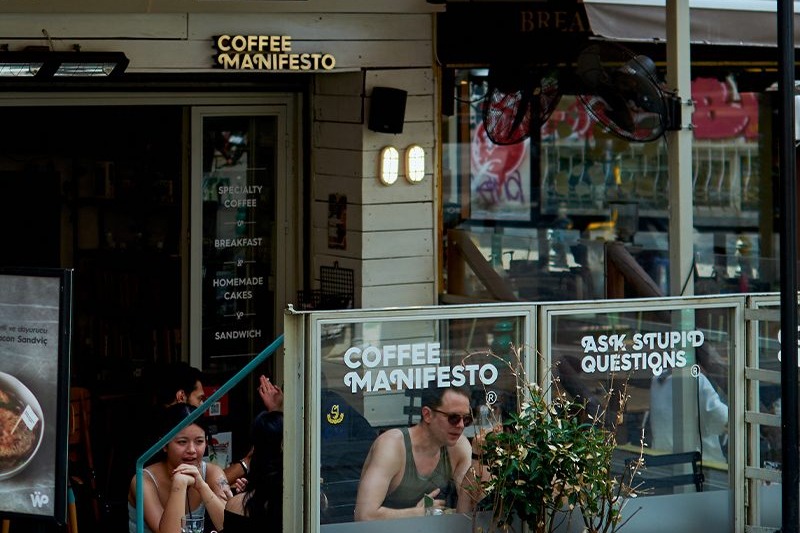 The Coffee Manifesto storefront in Moda, the name in gold letters above the door and etched on the glass windbreak, with customers seated on the pavement terrace