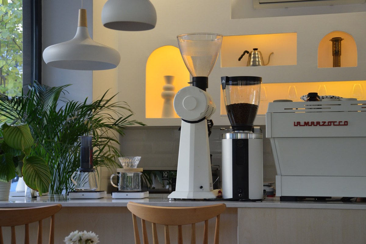 The bright bar at Good Coffee Roasting Co. in Kadıköy, white grinders and a La Marzocco machine in front of glowing arched shelves, with plants and wooden chairs