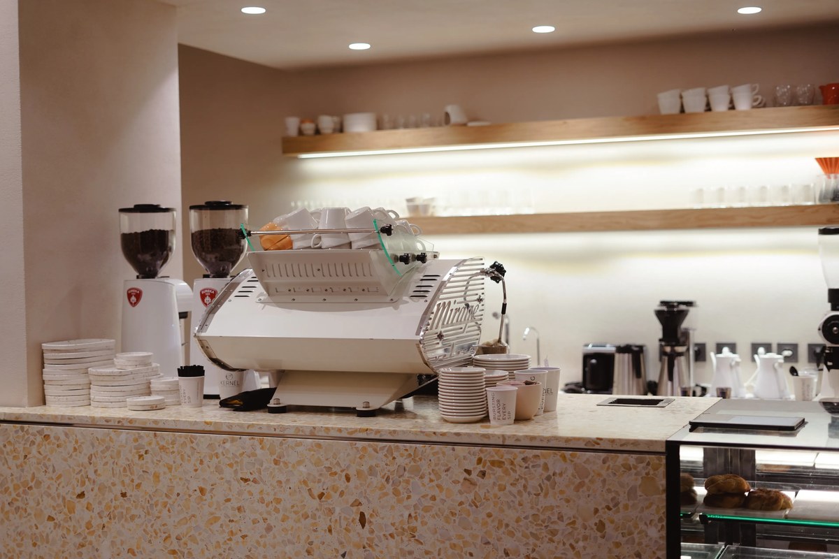 The terrazzo topped bar at Kernel Coffee in Suadiye, white grinders and a chrome and white espresso machine below lit wooden shelves of cups