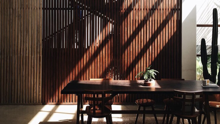 A wood slatted interior wall with dramatic afternoon light falling across a long communal table and chairs