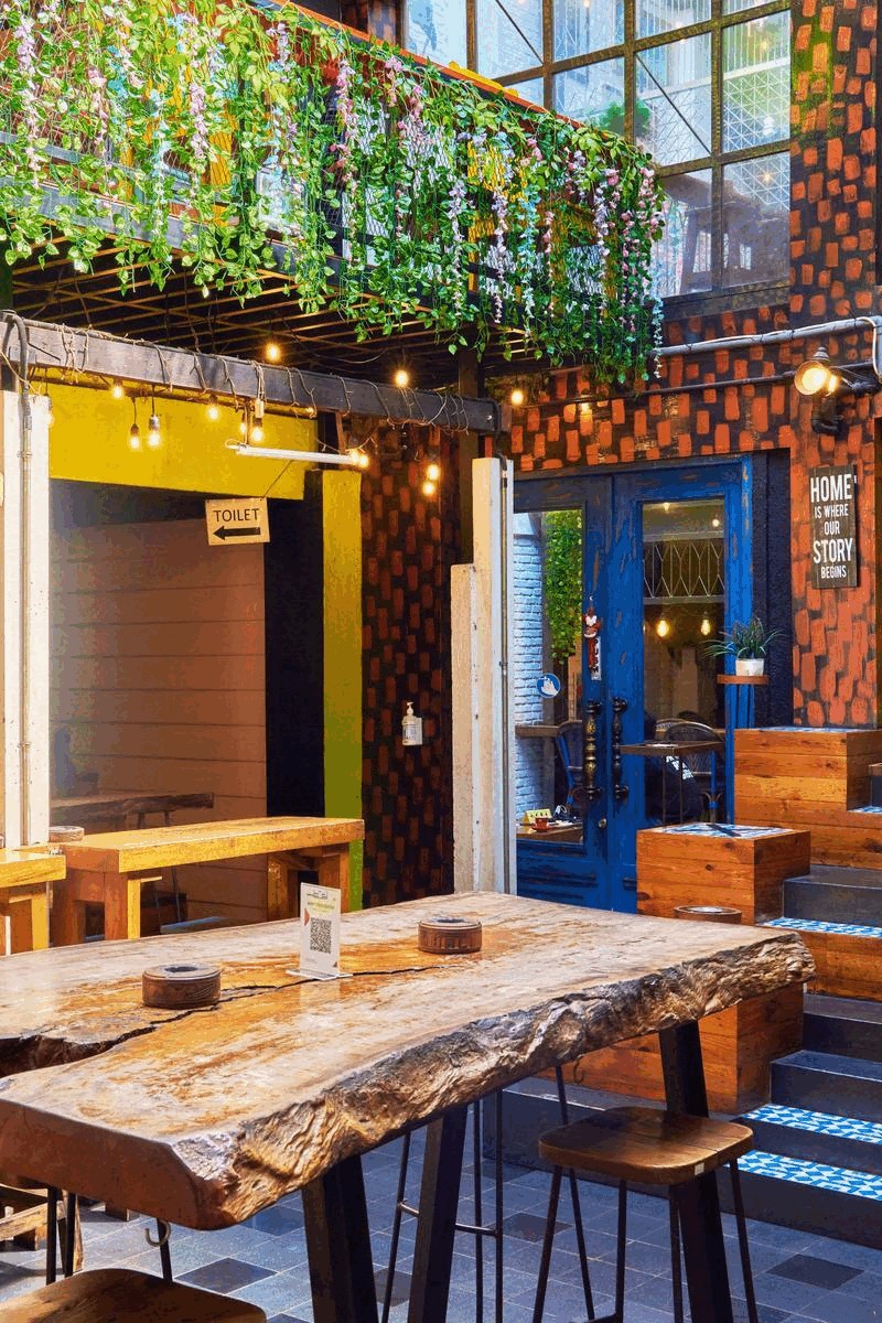 A plant draped courtyard with exposed brick walls, string lights, a live edge wood table and a blue door reading Home Is Where Our Story Begins