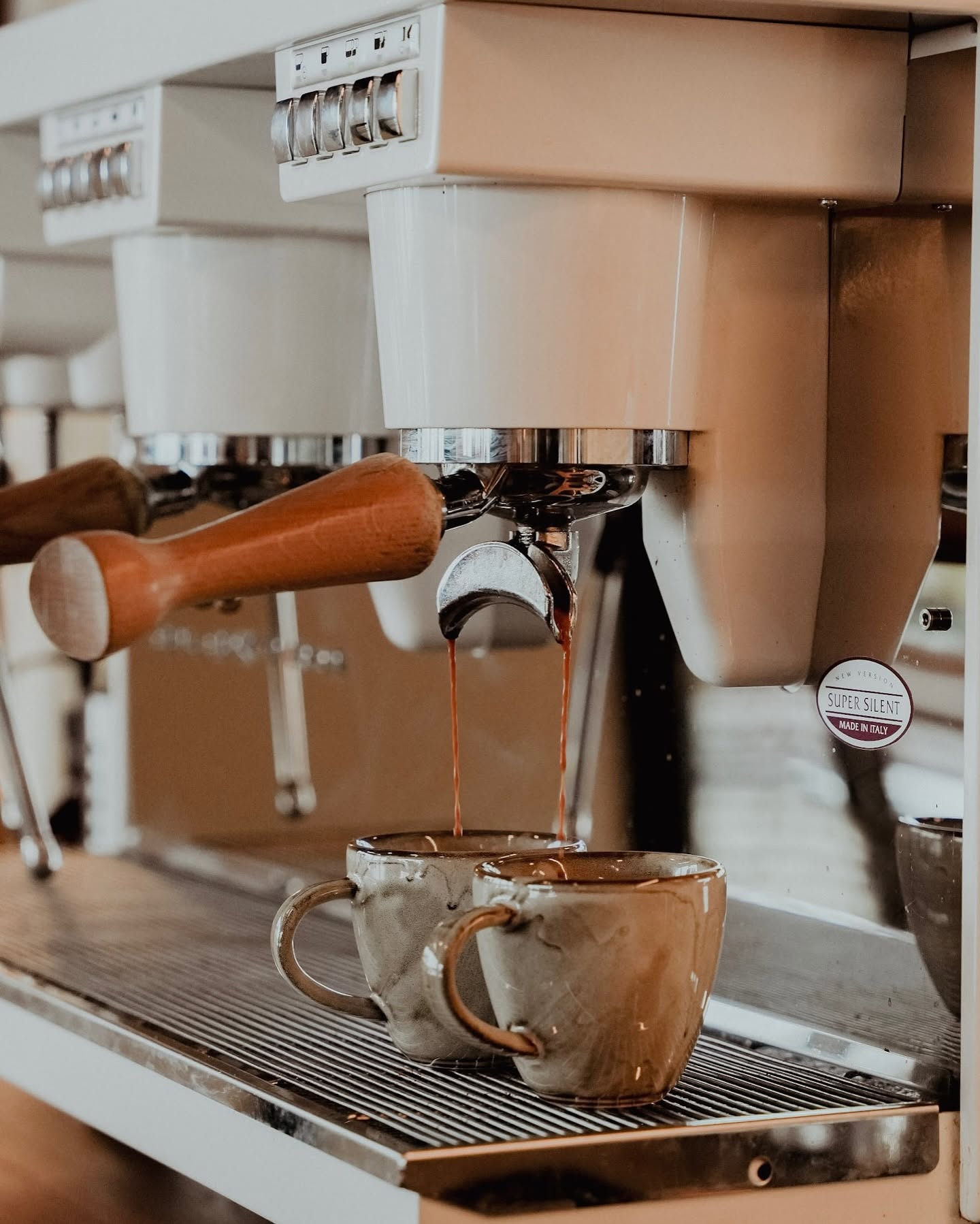 An espresso machine pulling a double shot into two mottled ceramic cups, with a wooden-handled portafilter resting on the group alongside
