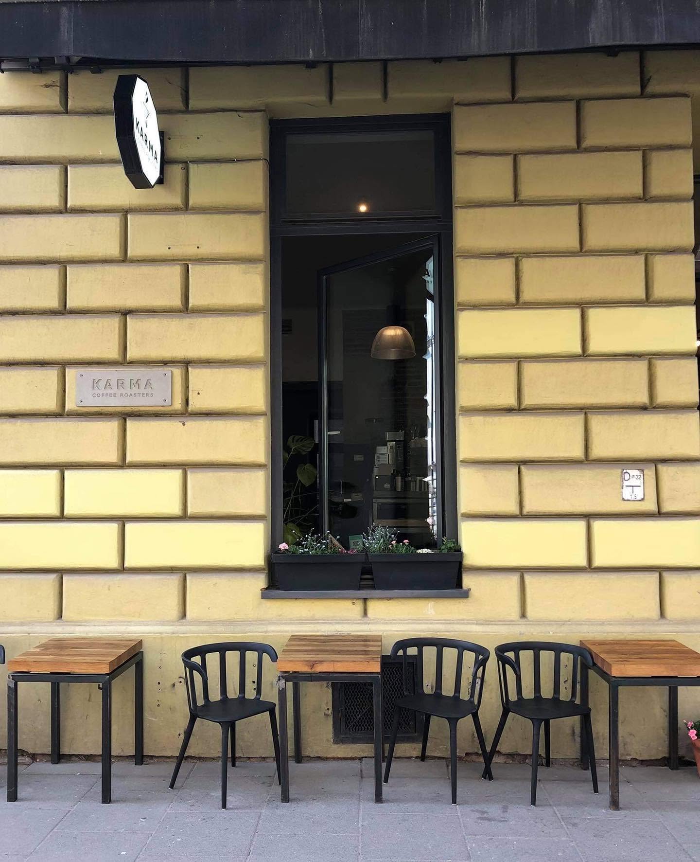 Karma Coffee Roasters' yellow ashlar frontage with an engraved KARMA COFFEE ROASTERS plaque, a hanging sign, a tall window with flower boxes, and wooden pavement tables with black chairs