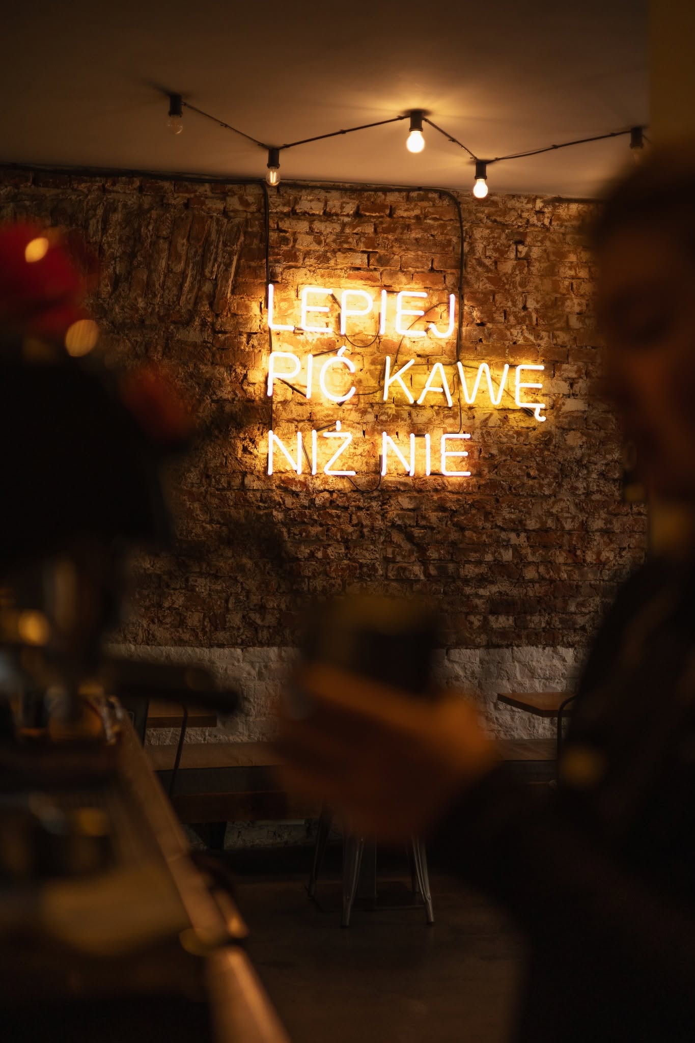 A warm neon sign reading LEPIEJ PIĆ KAWĘ NIŻ NIE glowing on an exposed brick wall under string lights, with cafe-goers softly blurred in the foreground