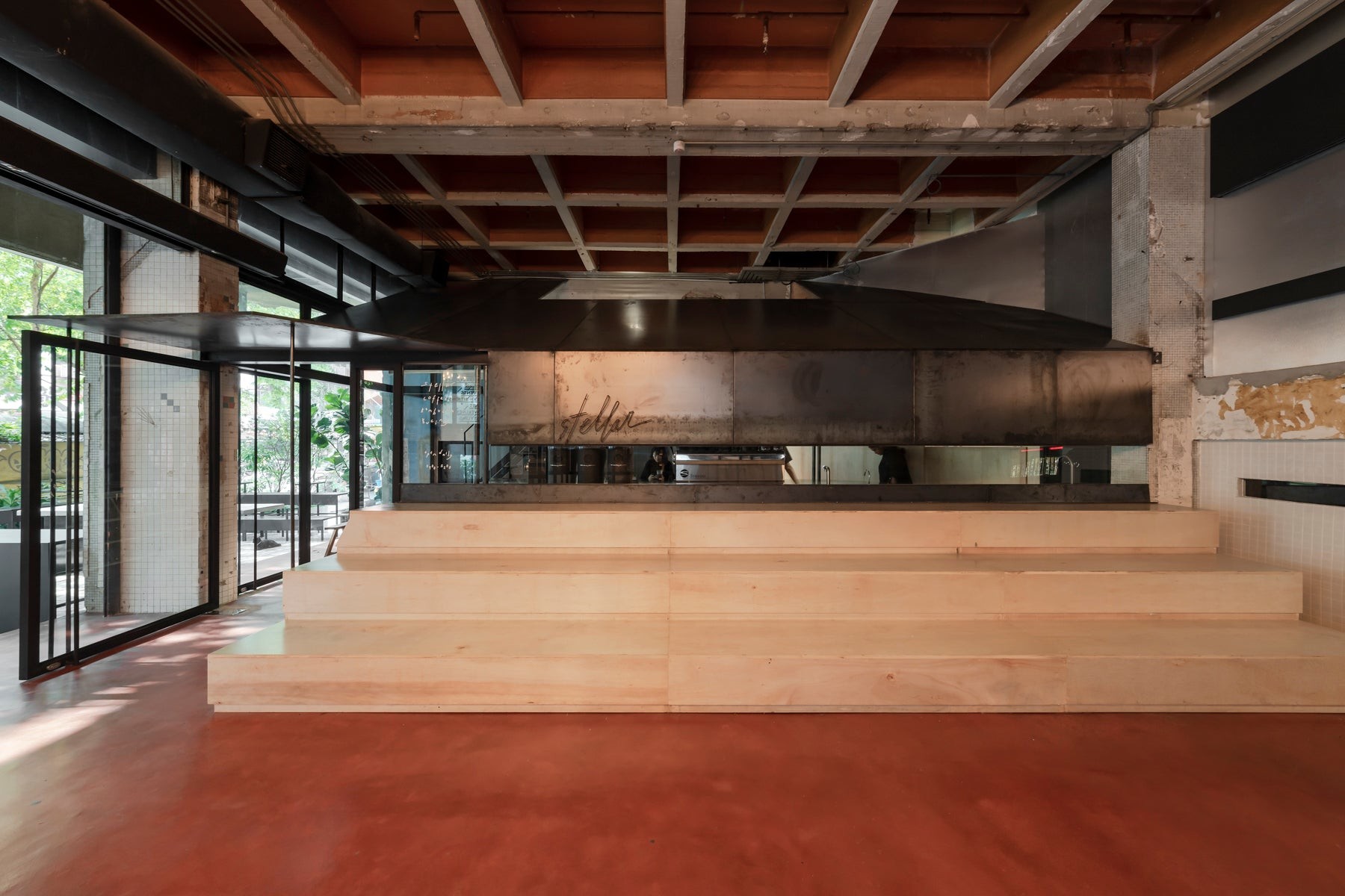 Pale plywood steps leading up to a steel hooded counter with the Stellar script on its front, inside a high ceilinged room with a red concrete floor and a leafy courtyard beyond the glass doors