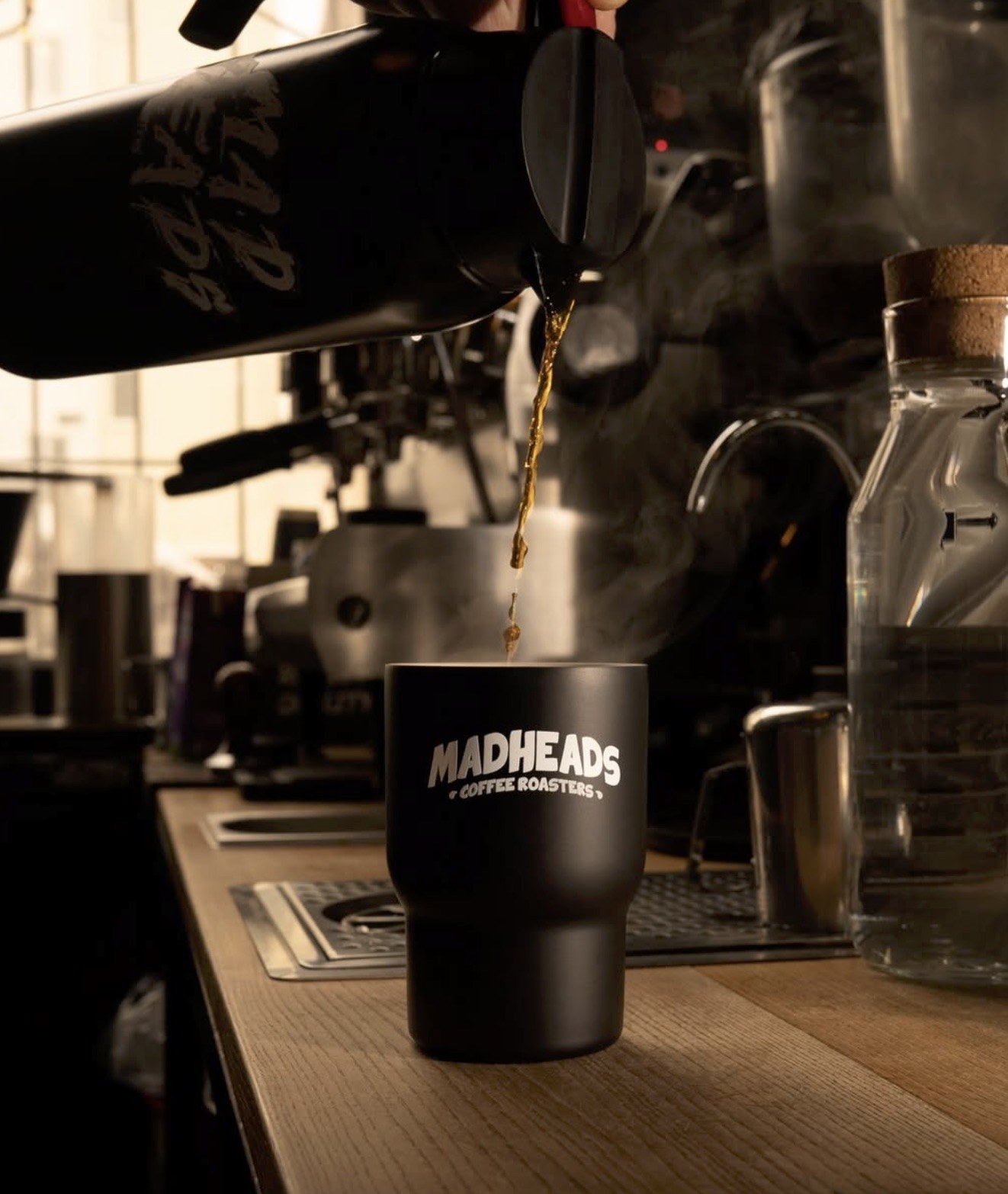 Coffee being poured from a branded Mad Heads carafe into a black Mad Heads Coffee Roasters cup