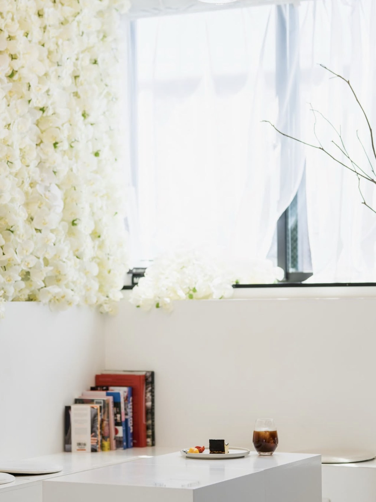 An iced coffee and a slice of cake on a white marble table, set against a wall of white flowers and a sheer curtained window