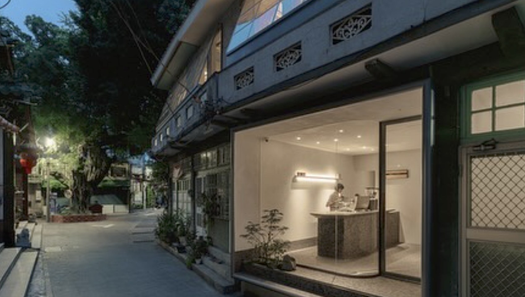 The machiya facade at dusk seen from the street, glass storefront lit from within with a barista working at the stone counter
