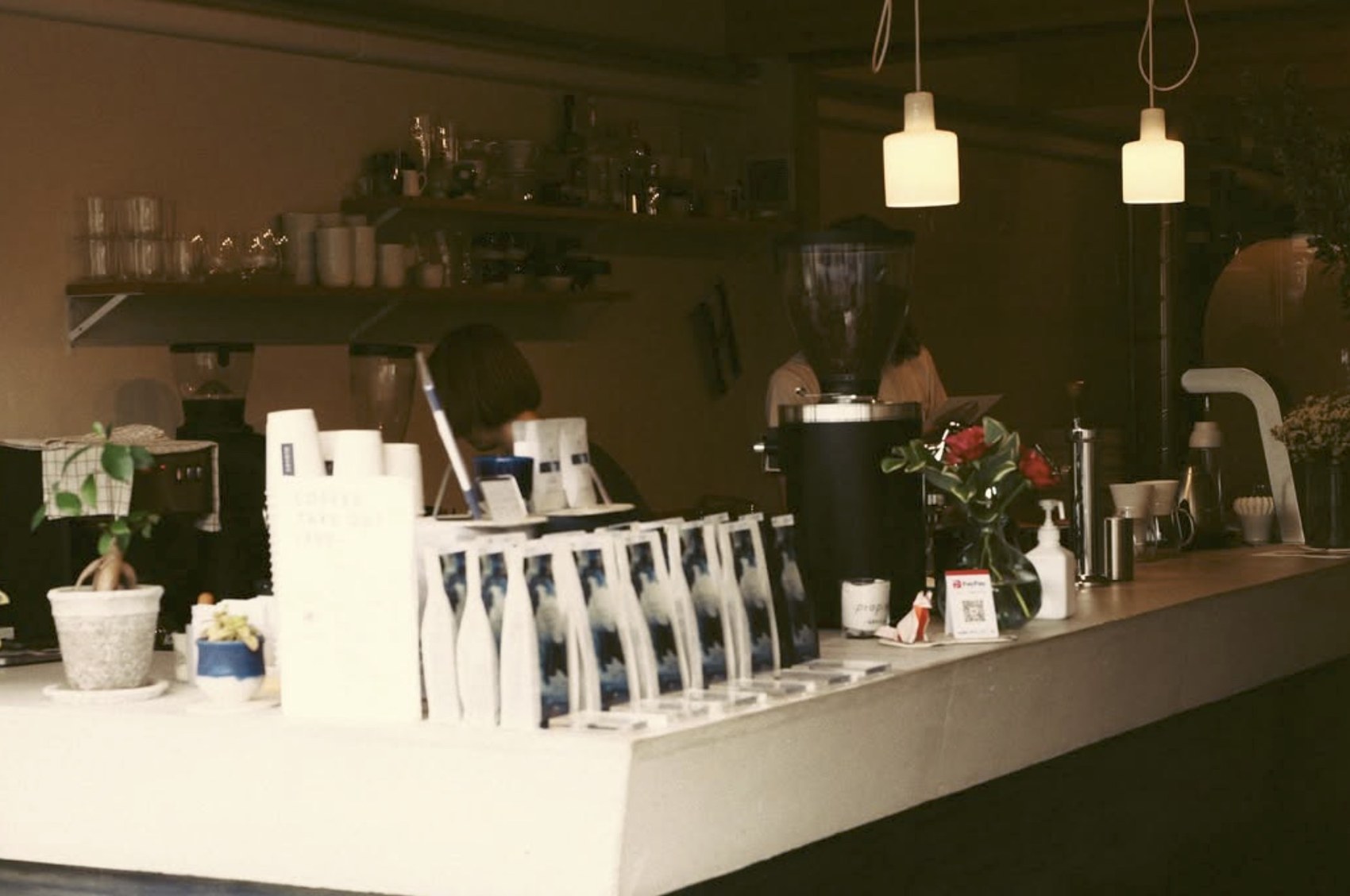 A warmly lit counter with retail bean bags lined up beside a grinder and a vase of roses, staff working at the bar