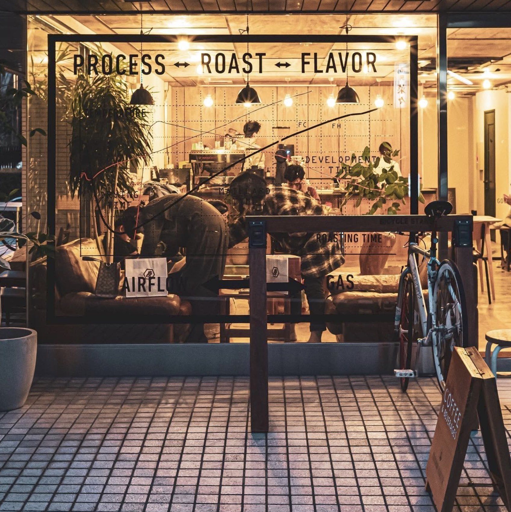 The glass storefront at night with a roasting process diagram lettered on the window, a bicycle parked outside and staff working the counter within