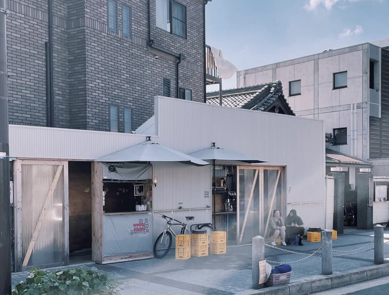 The corrugated metal storefront at dusk with two market umbrellas, crate seating, a bicycle parked out front and customers seated outside