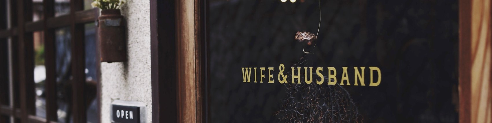 Brass Wife & Husband lettering on a dark storefront door, a bundle of dried flowers hanging above and an OPEN sign beside the entrance