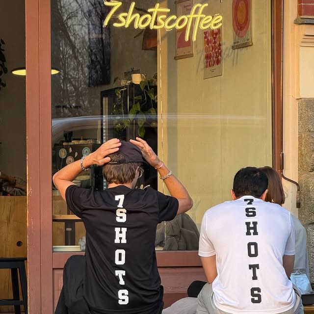 Two people with their backs turned, wearing matching 7 Shots branded t shirts, sitting outside the shop's window beneath its glowing 7shotscoffee sign