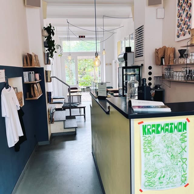 A narrow cafe hallway with a dark blue wall lined with bagged coffee and a hanging t shirt, leading past a wood counter with a coffee grinder to a bright doorway opening onto a garden