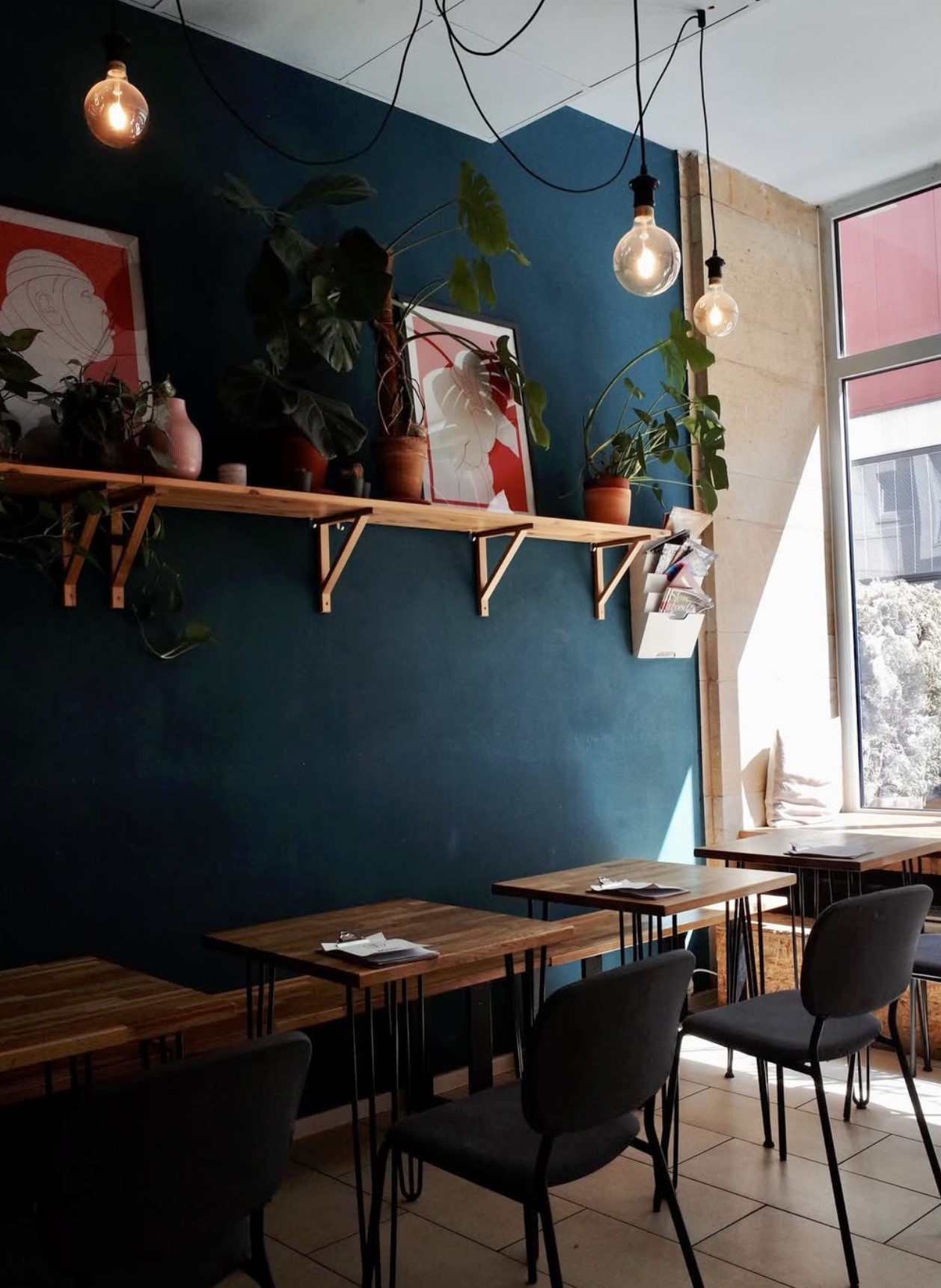 A dark blue dining wall at Bohemian Kids Café hung with framed art and lined with a wood shelf of plants, hairpin leg tables set below