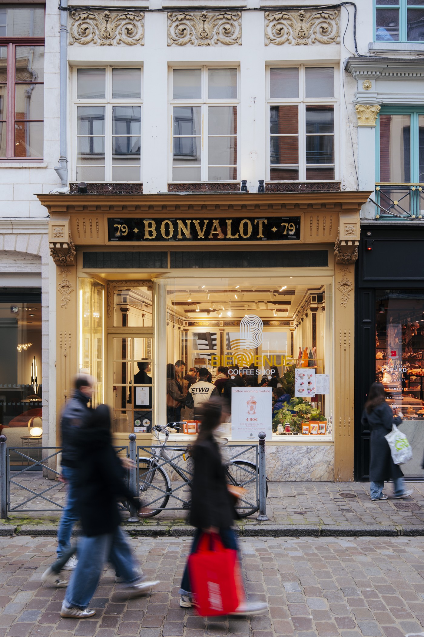 The gilded historic storefront on rue Esquermoise with the Bienvenue Coffee Shop sign lit in the window as blurred pedestrians pass by